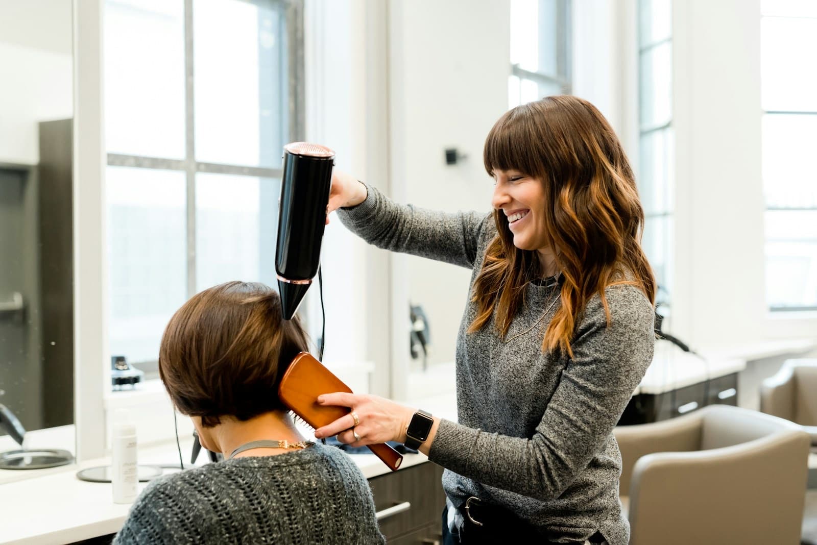A hairdresser laughing as she dries a client's hair in a bright salon